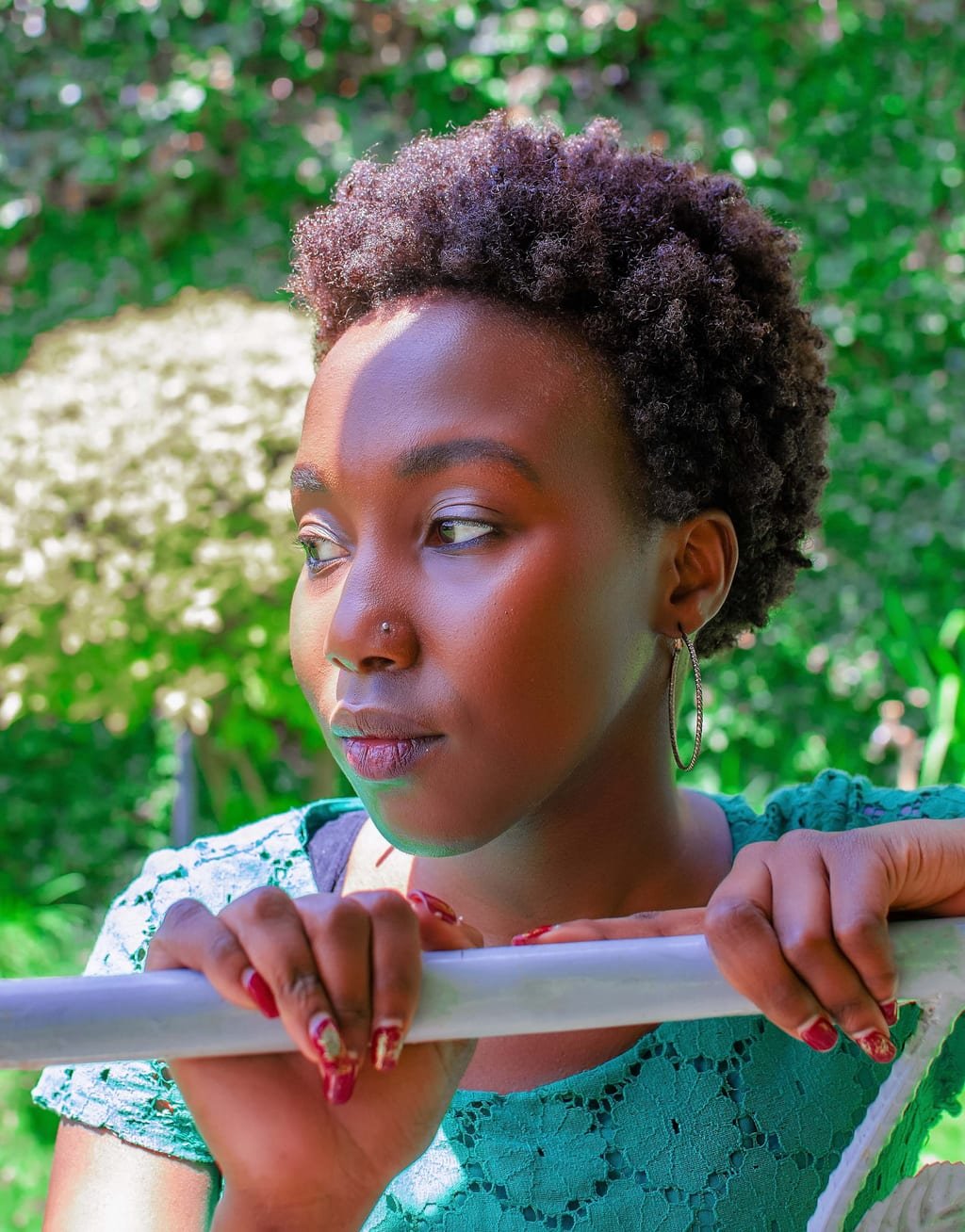 Kim leaning on a white railing in a green lace top, looking off to the side, in a sunlit garden
